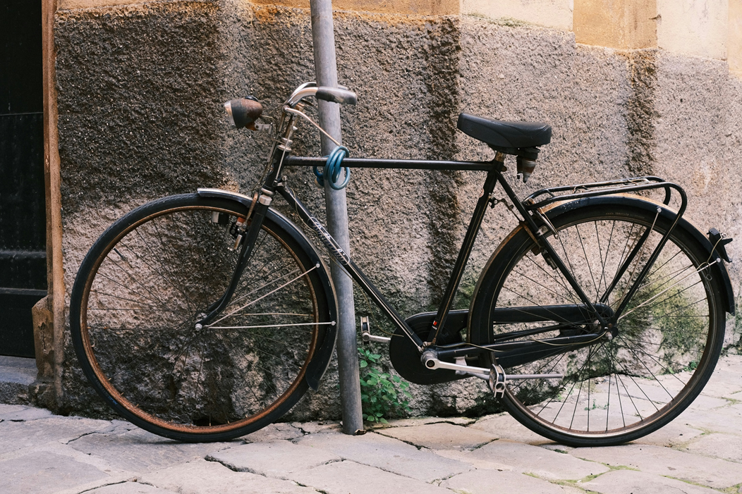 A touring/cruiser-style bicycle is propped against a steel pole, to which it is secured by a looped steel cable lock. A rough-surfaced exterior wall is in the background. Image credit: Indre B/Unsplash