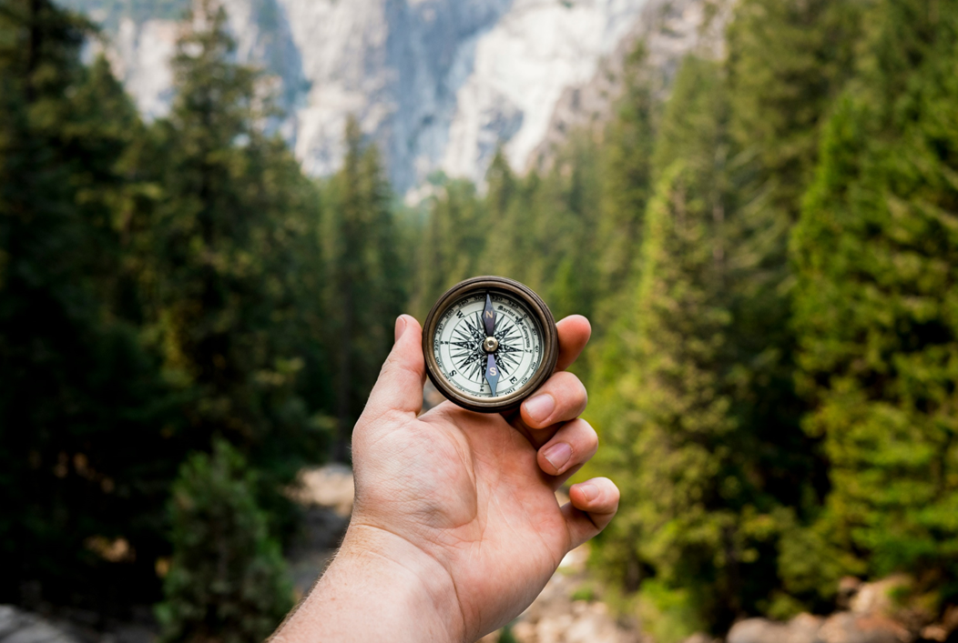 A person’s hand holds up a brass compass showing the face, the north/south needle pointing straight ahead in the picture’s field of orientation. Out of focus in the background are tall conifer trees and rocky mountain faces. Image credit: Jamie Street/Unsplash