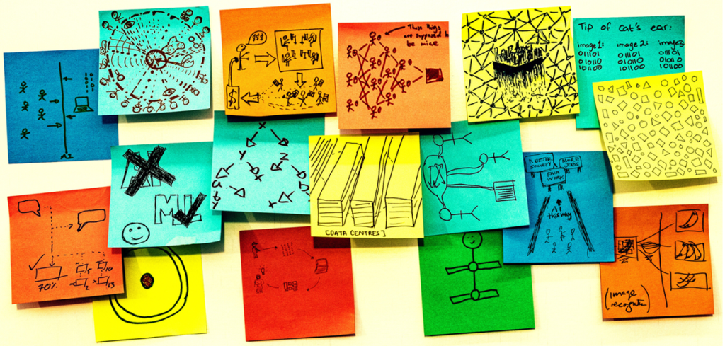 Seventeen multicoloured post-it notes are roughly positioned in a strip shape on a white board. Each one of them has a hand drawn sketch in pen on them, answering the prompt on one of the post-it notes "AI is...." The sketches are all very different, some are patterns representing data, some are cartoons, some show drawings of things like data centres, or stick figure drawings of the people involved. Image credit: Rick Payne and team / Better Images of AI / Ai is... Banner / CC-BY 4.0