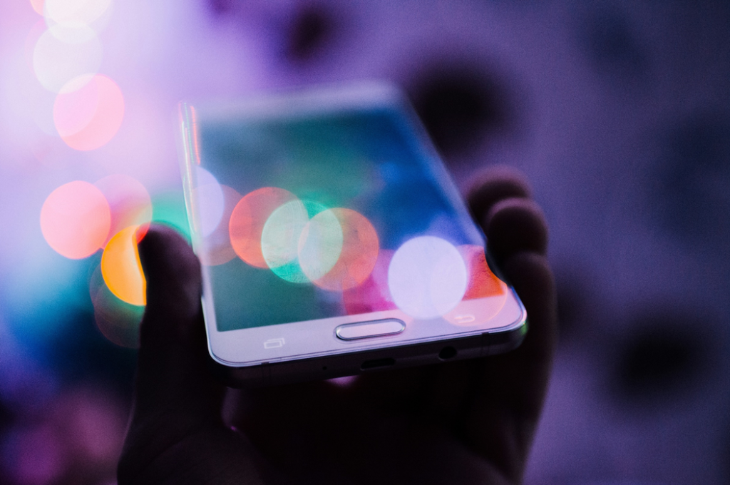 Artistically blurred, selective-focus photograph of a hand holding a smartphone up with blurred colored lights superimposed. Image credit: Rodion Kutsaiev/Unsplash