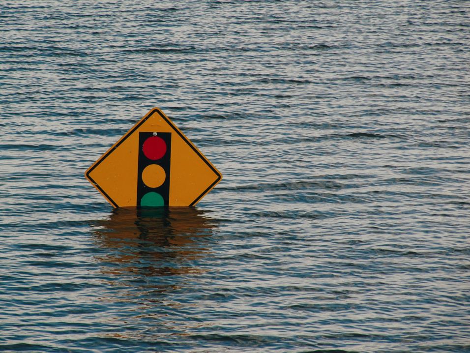 Photograph of a yellow traffic sign for “stop light ahead” standing in flood water that has reach the bottom corner of the sign. Image credit: Kelly Sikkema/Unsplash