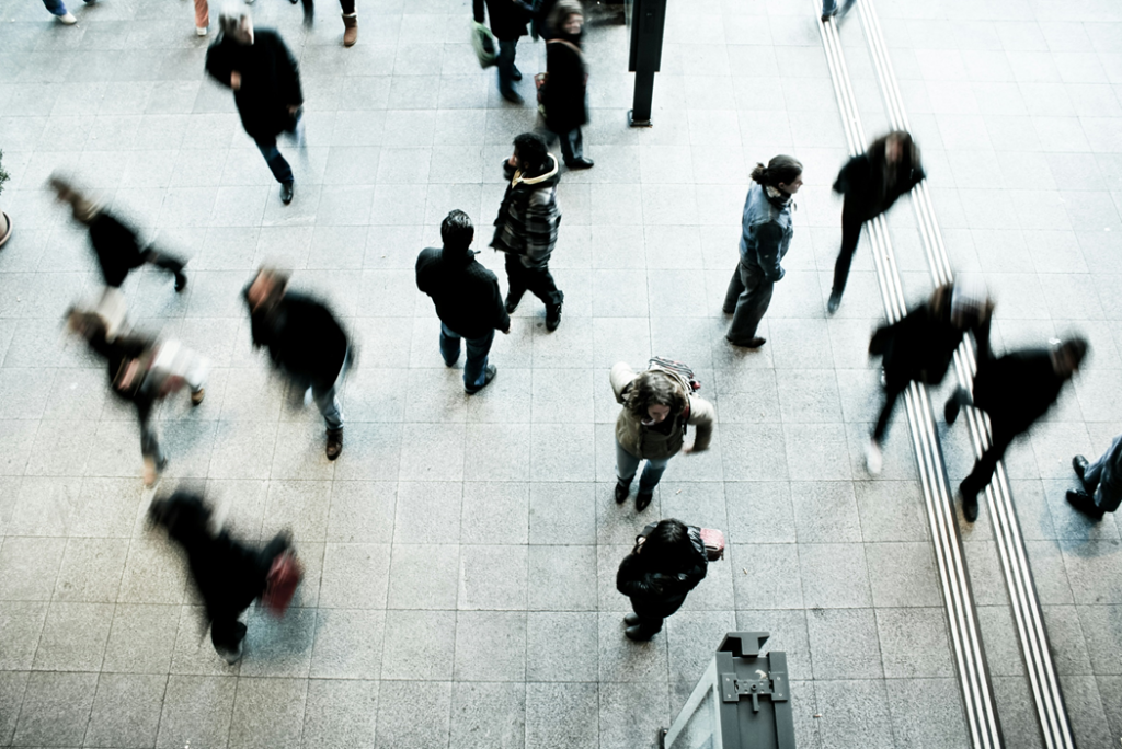 Photograph, taken from above/high angle, showing pedestrians, some of them motion-blurred, in a public square or train station. Image credit: Timon Studler/Unsplash