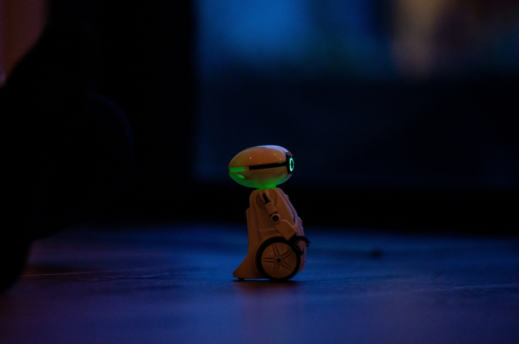 A small toy robot with wheels and glowing eyes, viewed in profile against a dark, indistinct background. Image credit: Jochen van Wylick/Unsplash