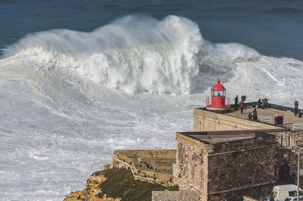 Large wave breaking in the bay of Nazare, Portugal, with a stone-masonry building and lighthouse on a promontory in the foreground. Luis Ascenso from Lisbon, Portugal - Praia do Norte, Nazaré, Portugal, CC BY 2.0, https://commons.wikimedia.org/w/index.php?curid=57820621