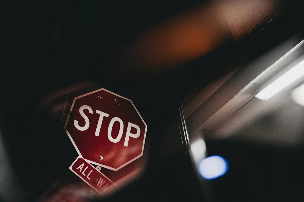 Stop sign with “all way” sign hanging crooked underneath, against a dark background and fluorescent lighting. Image credit: Jake Allen/Unsplash