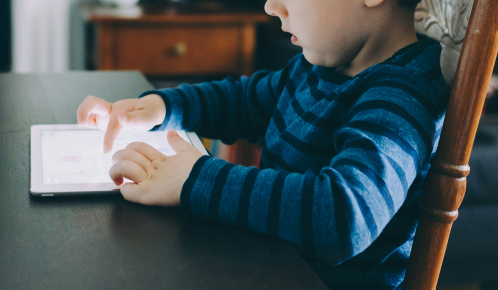 A small child sits at a table, absorbed in a task or game on a glowing computer tablet. Image credit: Kelly Sikkema/Unsplash