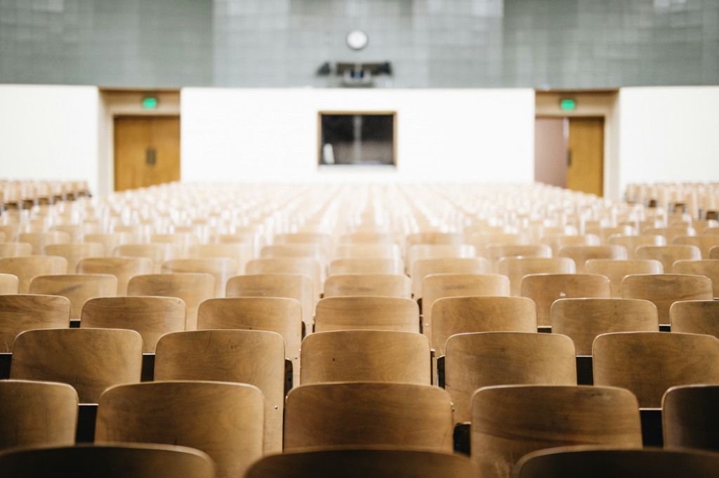 Photograph of rows of wood chairs/desks in a college auditorium-style classroom. Nathan Dumlao/Unsplash