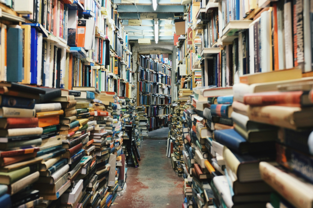 Photograph down a narrow, book-crowded corridor in the stacks of a library, with semi-orderly heaps of books in the foreground and shelved books reaching to the ceiling. Image credit: Glen Noble/Unsplash