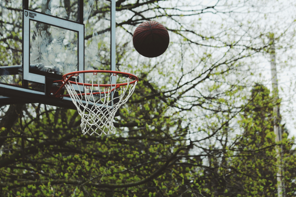 Basketball in mid-flight just above the rim of an outdoor basketball goal, with trees in the background. Image credit: Noah Silliman/Unsplash