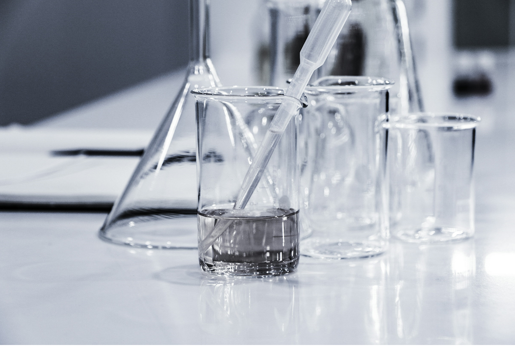 Photograph of laboratory glassware, including graduated cylinders, Erlenmeyer flasks, and pipettes on a table or benchtop. Image credit: Hans Reniers/ Unsplash