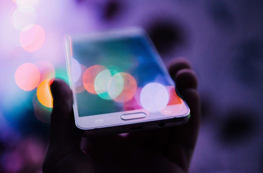 A hand holds up a smartphone against a blurred background of colorful lights. Image credit: Rodion Kutsaiev/Unsplash