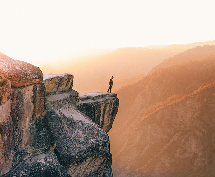 A person stands on the edge of a stony precipice, looking down into an enormous drop into a valley in a mountainous setting (photo cropped from original dimensions). Image credit: Leio McLaren/Unsplash