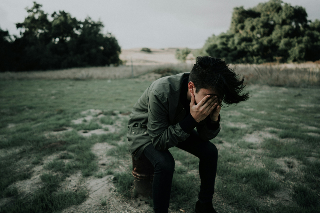 A young man sits in an outdoor clearing with trees in the background, with hands covering his face as if distraught. Image credit: Francisco Gonzalez/Unsplash