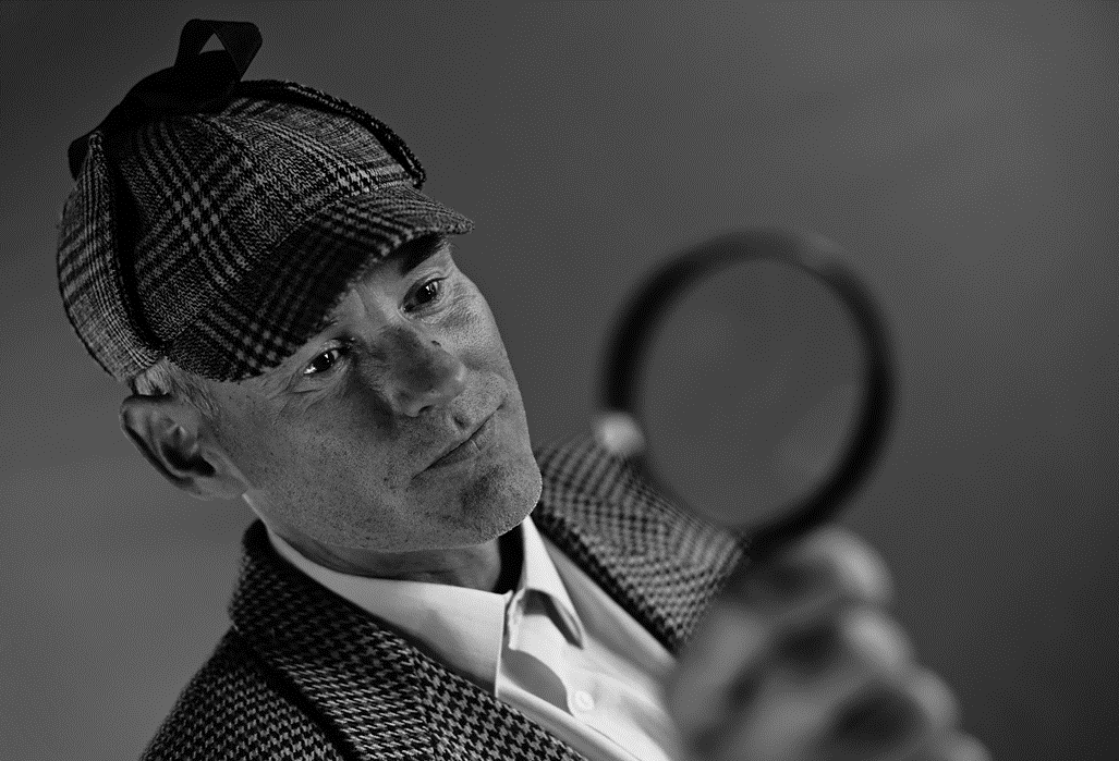 Black and white photo of a man wearing a Sherlock-Holmes-style tweed jacket and deerstalker cap holds up a magnifying glass, out of focus in the foreground. Image credit: Andres Siimon/Unsplash