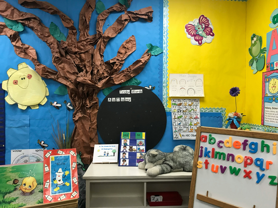 Photograph of an empty primary school classroom with a bookshelf, bulletin boards, colorful posters, magnetic alphabets, and stuffed animals. In the cener is large black disk with the words: “Tricky Words: AI is my”. Image credit: Monica Sedra/Unsplash