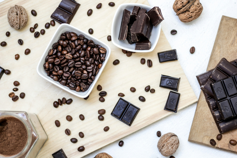 Photograph, taken of a white table top with cutting boards from above, showing chunks of chocolate, coffee beans both loose and in a dish, a small dish containing powdered cocoa, and unshelled walnuts. Image has been rotated 90 degress counterclockwise from original orientation. Image credit: NordWood Themes/Unsplash