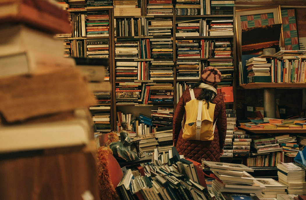 A person wearing a backpack, with back turned toward the camera’s perspective, looks at shelves of old books stacked flat on their sides, with disorderly heaps of books in the foreground. Image credit: Darwin Vegher/Unsplash