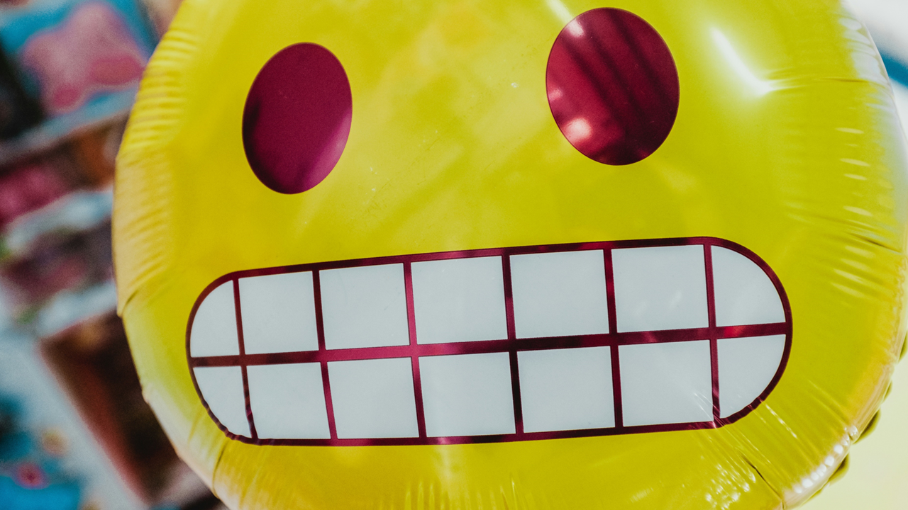 Photograph shows a balloon printed with the gritted-teeth emoji expressive of secondhand embarrassment. Image credit: Bernard Hermant/Unsplash