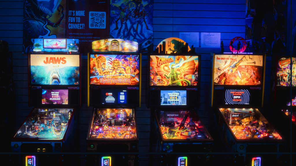 A row of four colorful, glowing pinball machines stand against the wall of dark arcade interior. Themes for each machine, from left to right: Jaws (the movie), Iron Maiden (the British heavy metal band), Godzilla, and You Only Live Twice (James Bond movie). Image credit: Jason Leung/Unsplash