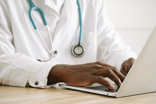 Image of a Black physician wearing a white coat and a stethoscope around his neck typing on a laptop